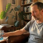A middle-aged man sits in a sunlit living room looking thoughtfully at a glass of water, appearing tired but resilient.
