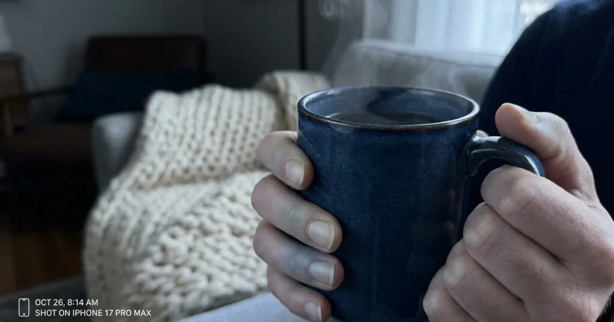 Close-up of pale hands clutching a steaming blue ceramic mug in a soft-lit living room.