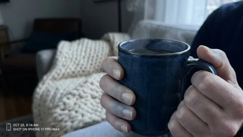 Close-up of pale hands clutching a steaming blue ceramic mug in a soft-lit living room.