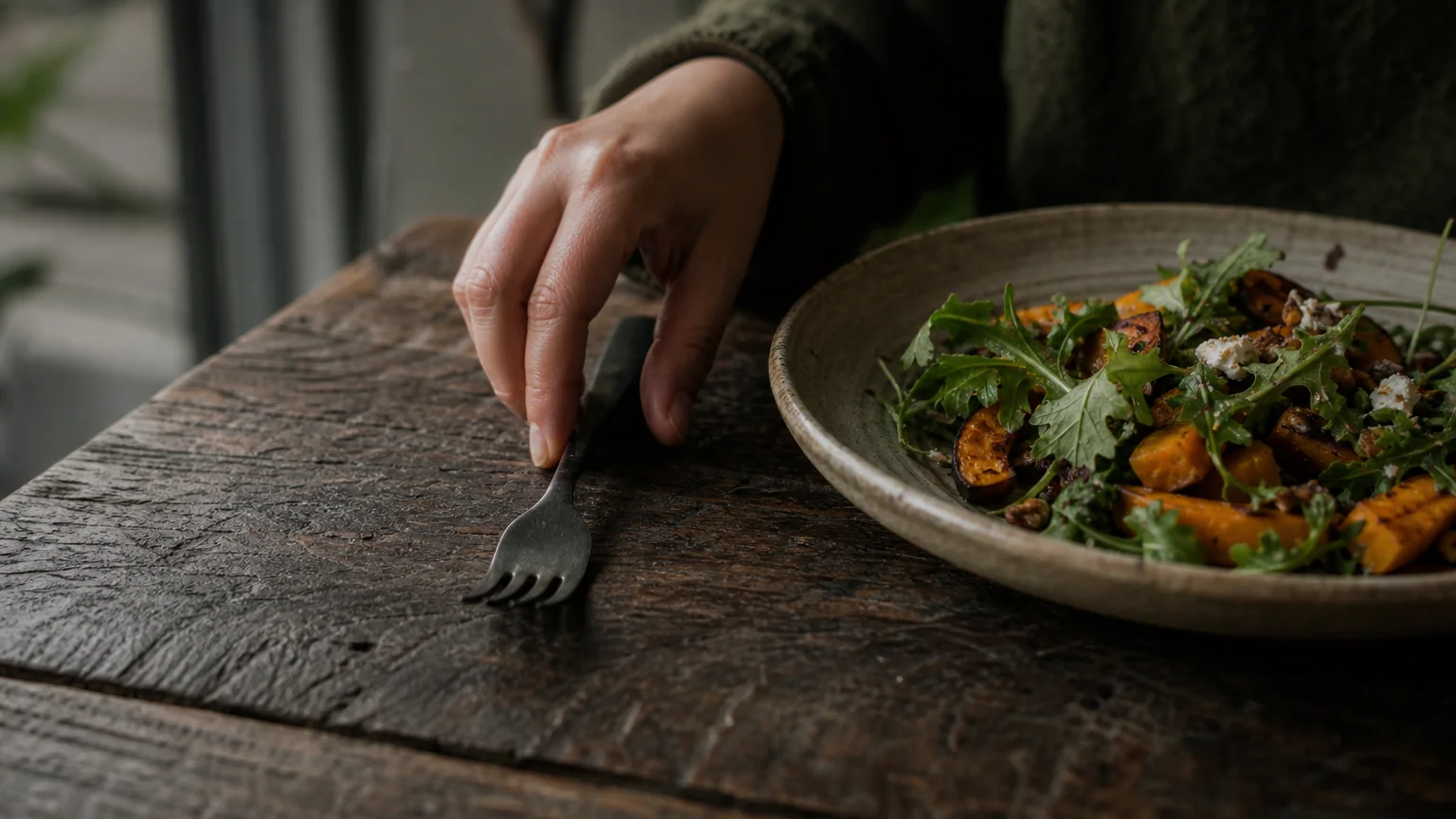 Close-up photo of a hand resting a ceramic fork on a rustic wooden table next to a plate of healthy food, emphasizing a pause in eating.