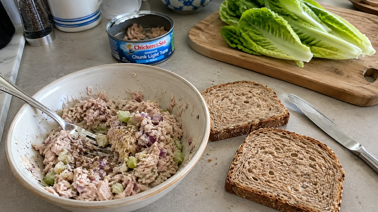 Tuna salad in a ceramic bowl next to two slices of whole grain bread, romaine lettuce, and an open can of tuna.