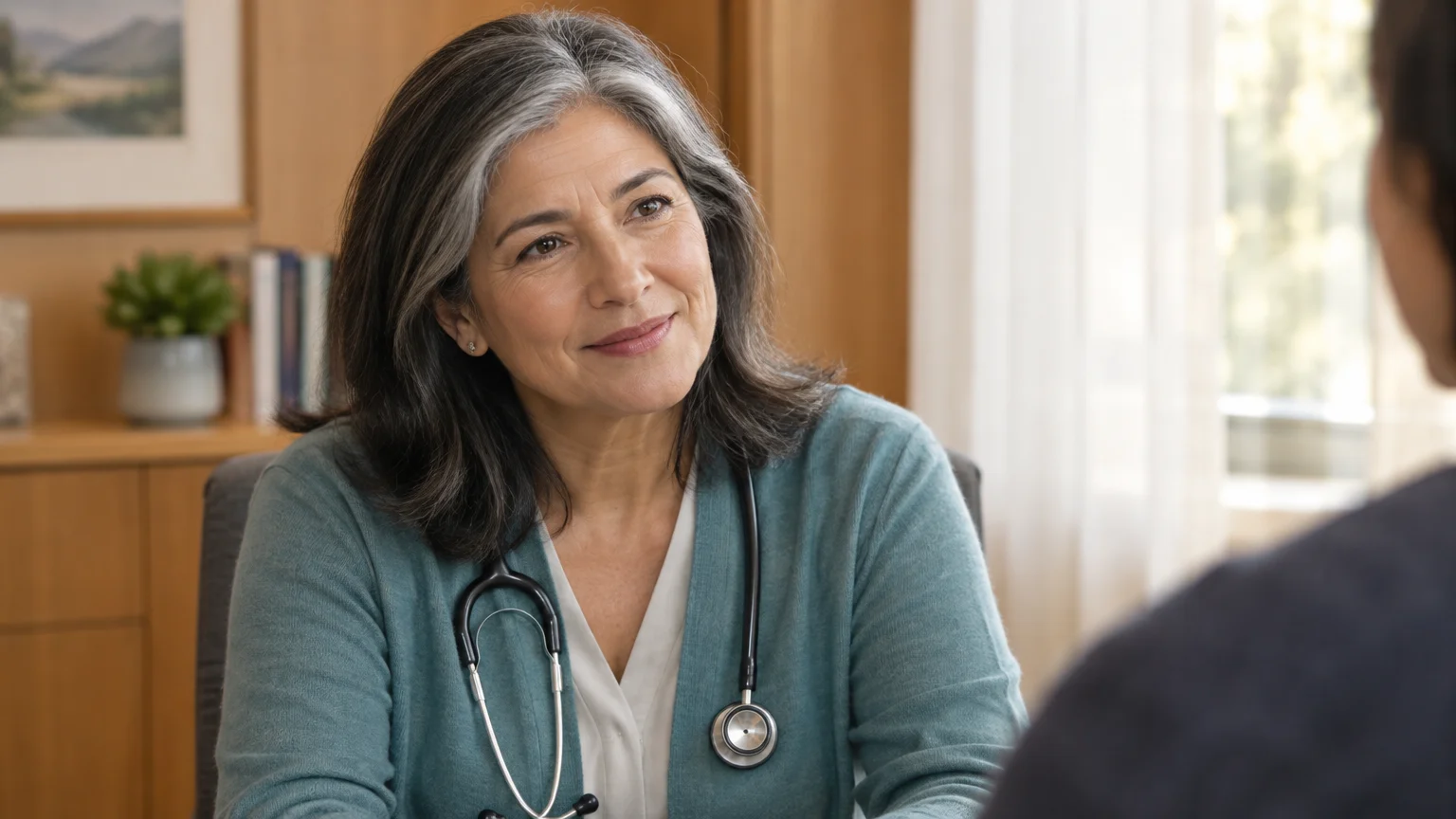 Smiling middle-aged female doctor with gray-streaked hair wearing a stethoscope and cardigan, talking with a patient.