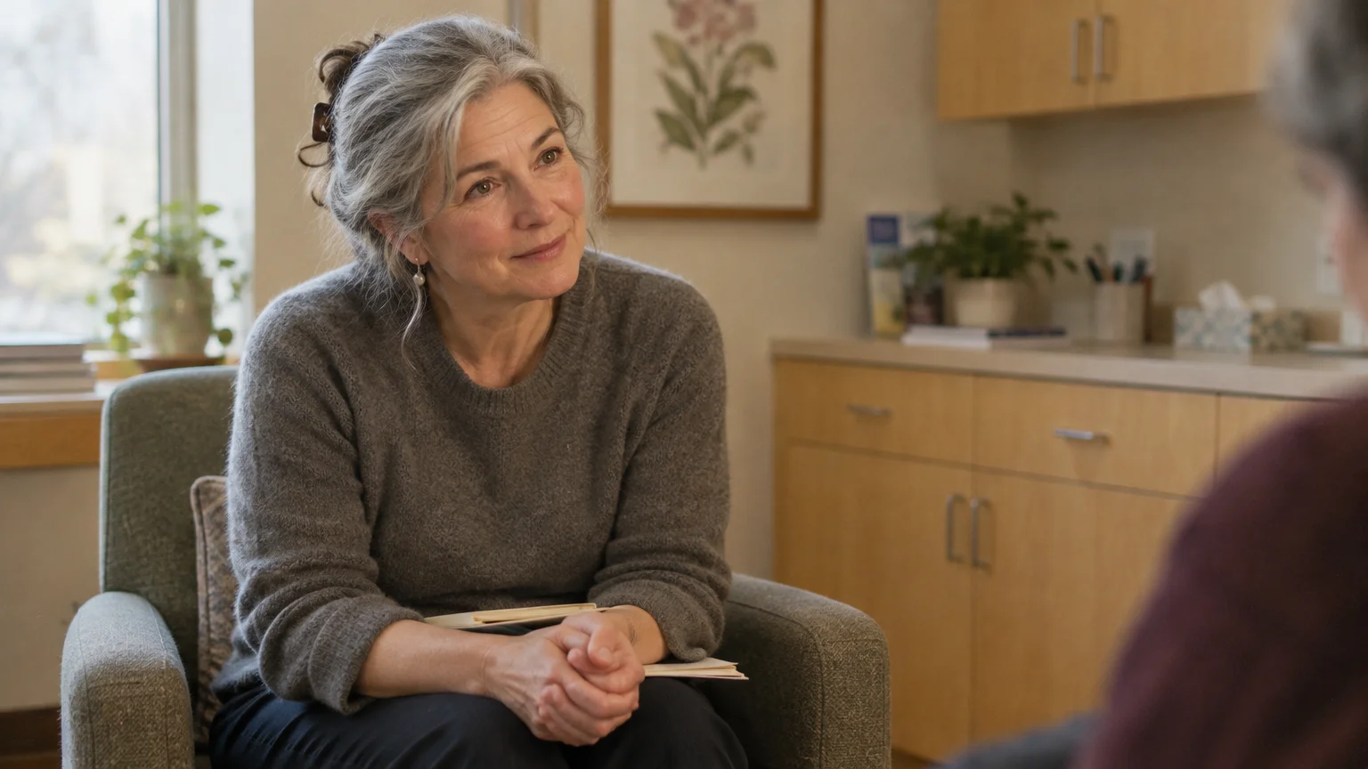 A smiling woman with gray hair sits in an armchair listening attentively in a clinical office setting.