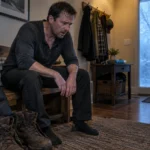 Exhausted, sweating man in dark clothes sitting on a wooden entryway bench next to work boots and a winter coat.